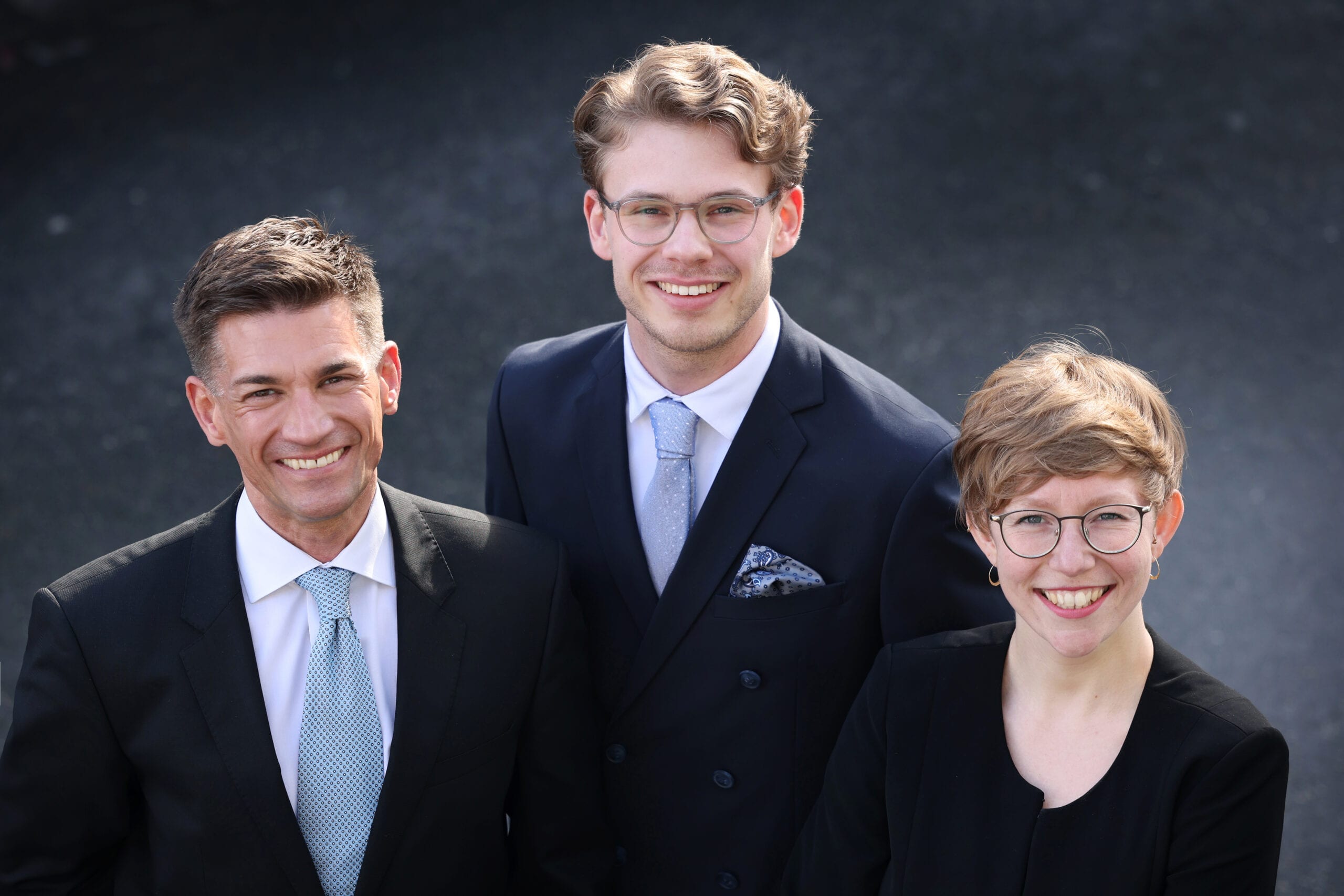 Auf dem Bild sind drei professionell gekleidete Personen abgebildet, die freundlich und zuversichtlich in die Kamera lächeln. Links steht Dr. André Latz in einem dunklen Anzug und heller Krawatte, in der Mitte Moritz Richarz in einem dunkelblauen, zweireihigen Anzug, und rechts Marie Geffroy, die einen eleganten schwarzen Anzug trägt. Ihre freundlichen Gesichtsausdrücke und die formelle Kleidung vermitteln Kompetenz und Professionalität.