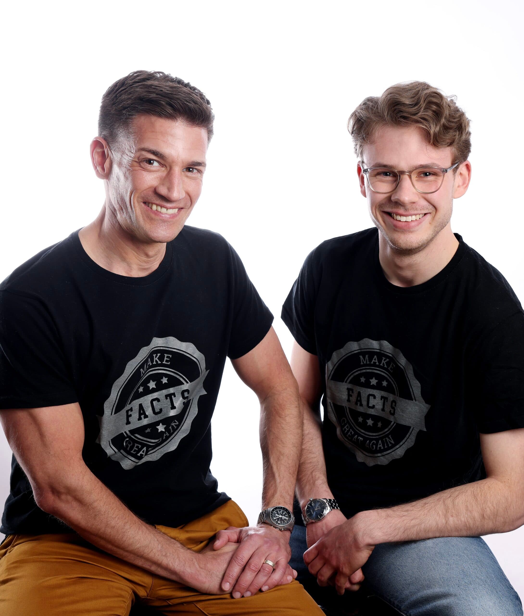 Foto von André Latz und Moritz Richarz André Latz (links) und Moritz Richarz (rechts) sitzen nebeneinander und lächeln in die Kamera. Beide tragen schwarze T-Shirts mit der Aufschrift „Make Facts Great Again“ in grauer Schrift auf einem runden Emblem. André Latz trägt braune Hosen, Moritz Richarz eine Jeans. Beide haben Uhren am Handgelenk. Der Hintergrund ist einfarbig weiß.