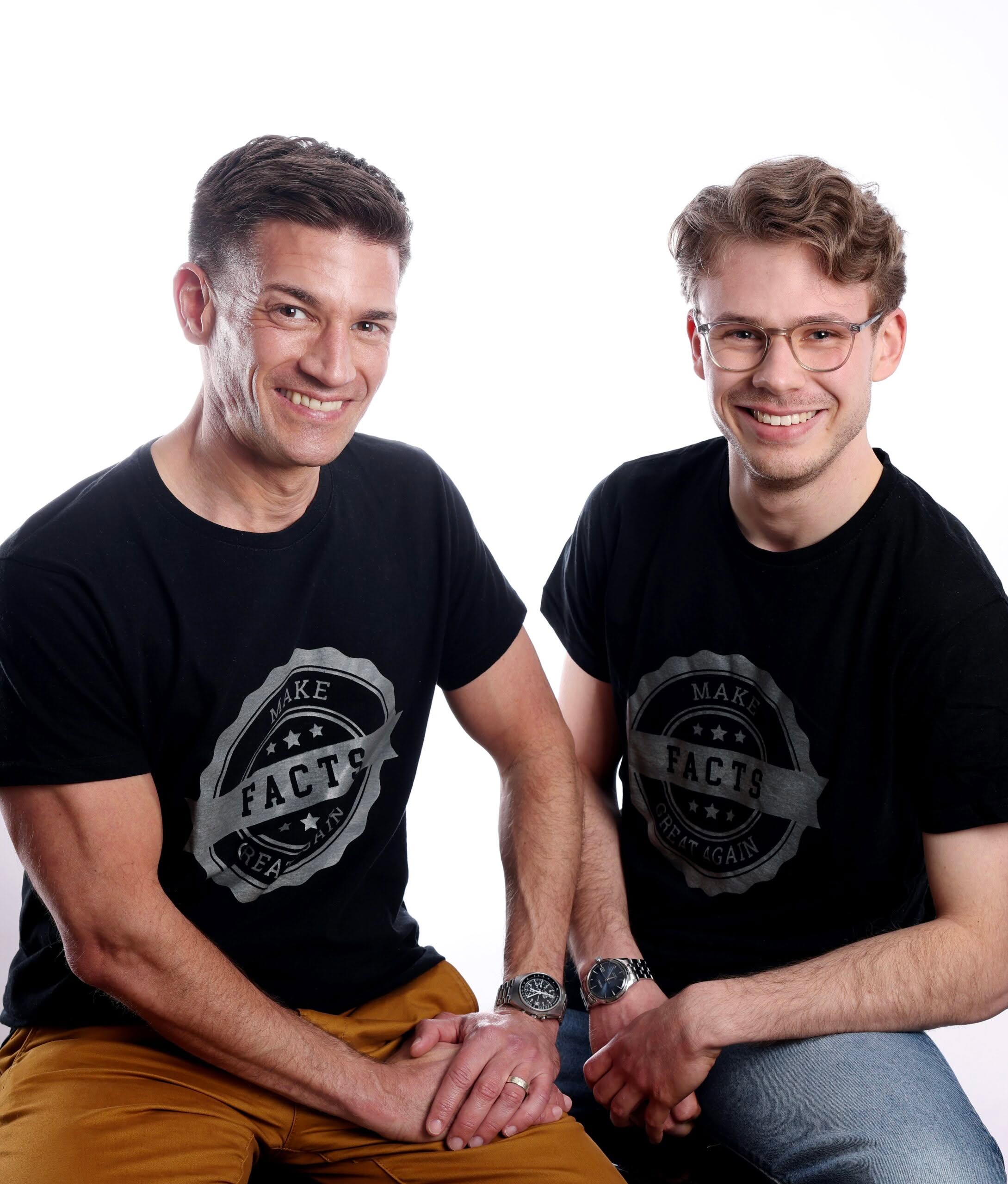Foto von André Latz und Moritz Richarz André Latz (links) und Moritz Richarz (rechts) sitzen nebeneinander und lächeln in die Kamera. Beide tragen schwarze T-Shirts mit der Aufschrift „Make Facts Great Again“ in grauer Schrift auf einem runden Emblem. André Latz trägt braune Hosen, Moritz Richarz eine Jeans. Beide haben Uhren am Handgelenk. Der Hintergrund ist einfarbig weiß.
