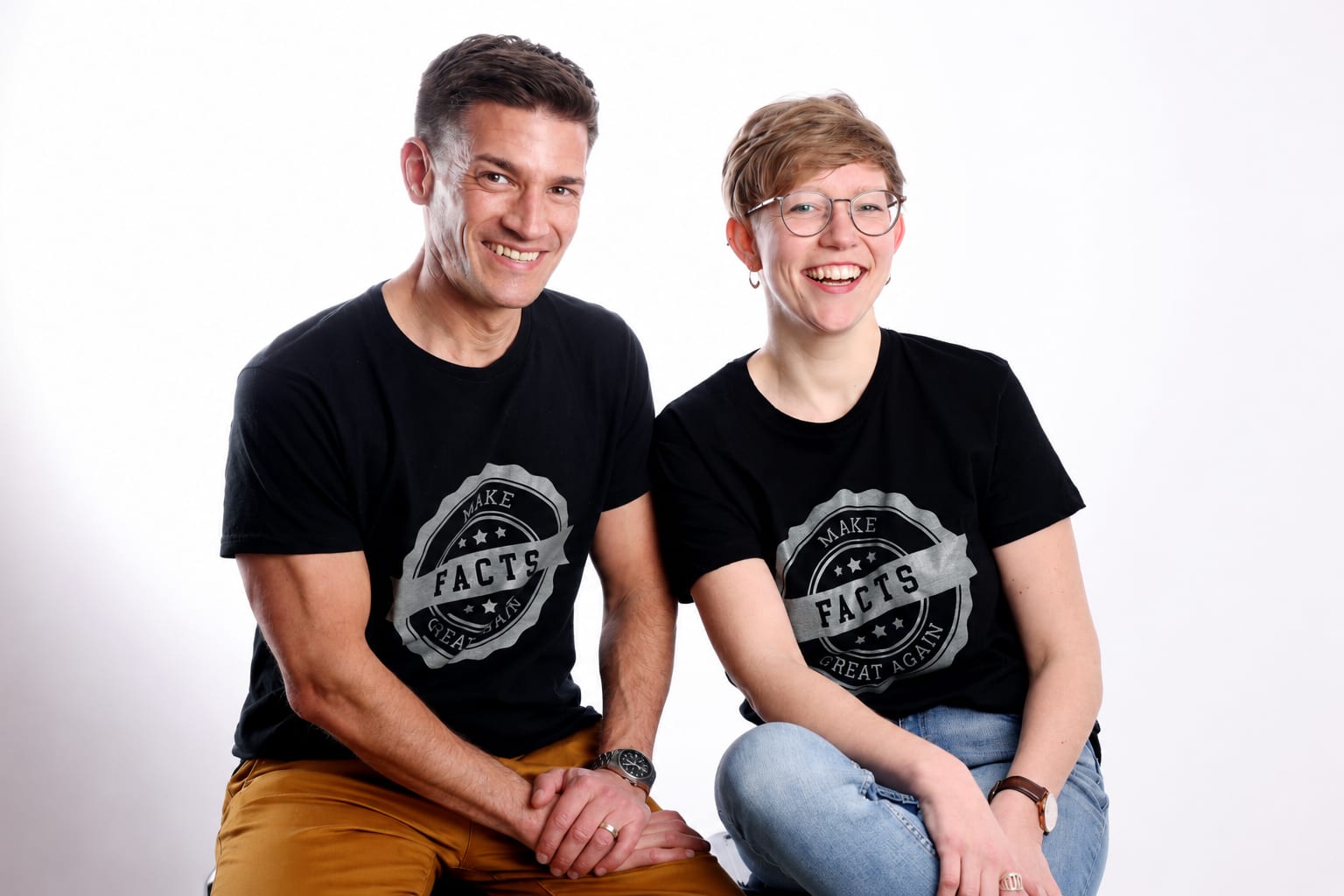 Facts-Shirt-Porträt Zwei lächelnde Personen sitzen nebeneinander in schwarzen T-Shirts mit der Aufschrift „Make Facts Great Again“.